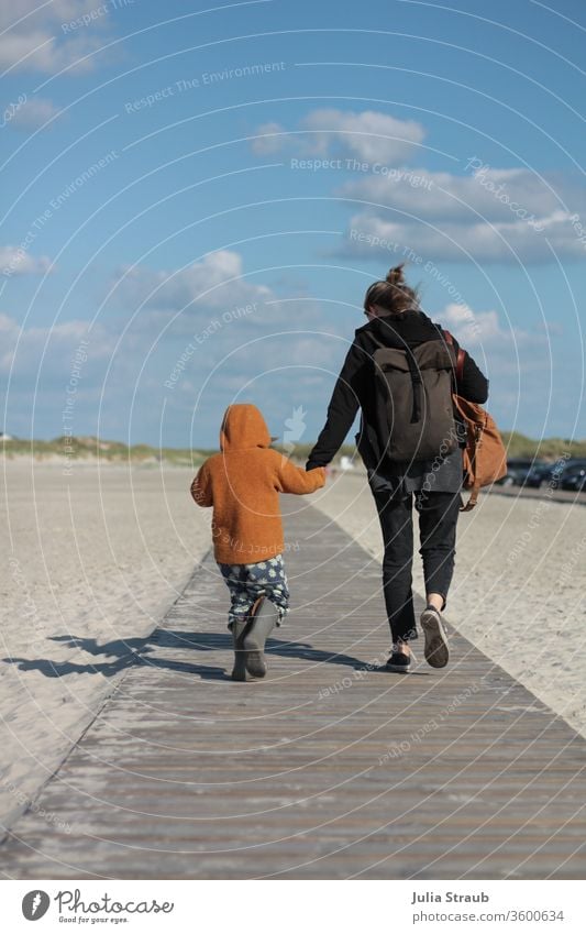 Frau und Kind laufen Händchen halten auf einem Holzweg zum Strand Sand weißer Sand Nordsee Dünen Dünengras Rucksack rucksacktourist Kleinkind wollwalk senfgelb