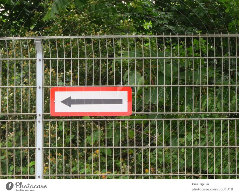 Gitterzaun mit Hinweisschild mit Pfeil, der nach links zeigt. Im Hintergrund grüne Sträucher und Bäume. Zaun Schilder & Markierungen Richtung Zeichen