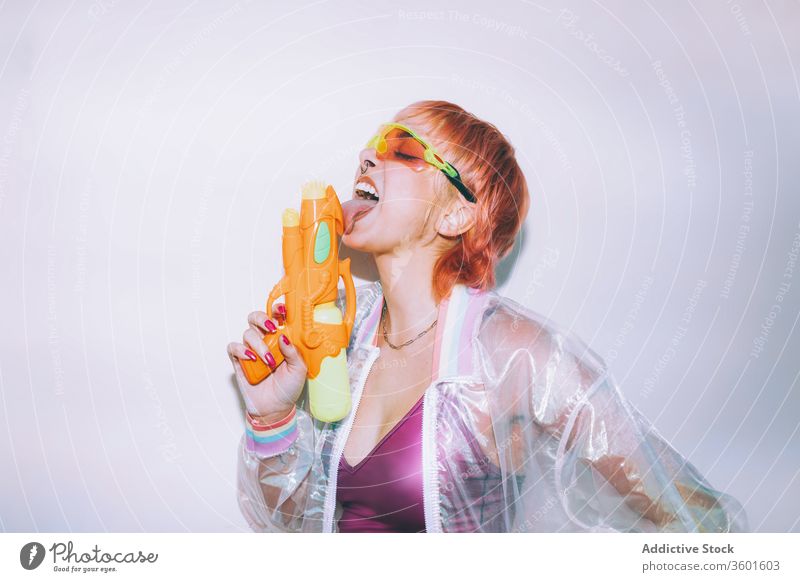Retro-futuristische Frau leckt Wasserpistole Pistole Stil Augen geschlossen lutschen jung Waffe Outfit retro Model gefärbtes Haar trendy Jacke Sonnenbrille