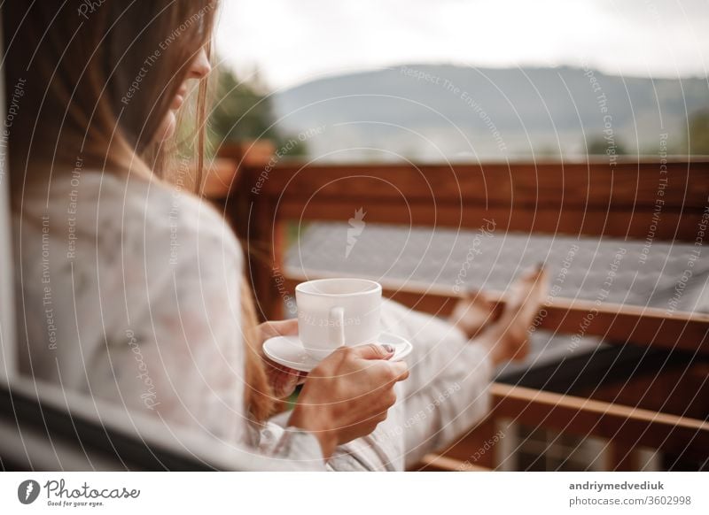 Junge Frau in stilvoller Nachtwäsche trinkt morgens gerne Kaffee oder Tee im Freien auf dem Balkon und schaut auf die Berge. Tasse heimwärts