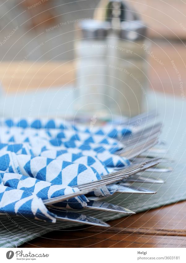 Mahlzeit! - Besteck - Messer und Gabel - eingewickelt in blau-weiße Servietten liegt auf einem Tisch, Salz- und Pfefferstreuer im Hintergrund Biergarten Lokal