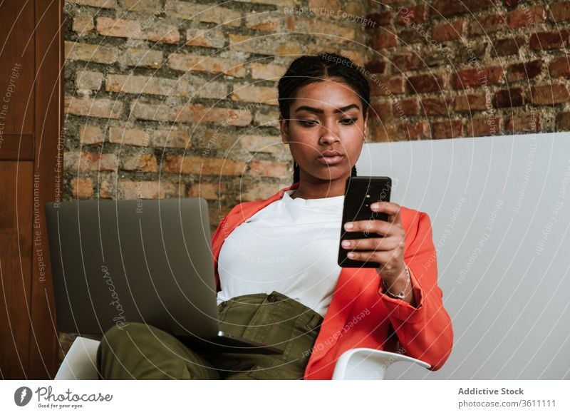 Moderne schwarze Frau mit Gadgets im Sessel Business Laptop heiter Smartphone online elegant Computer Loft Design Innenbereich freiberuflich modern Glück Gerät