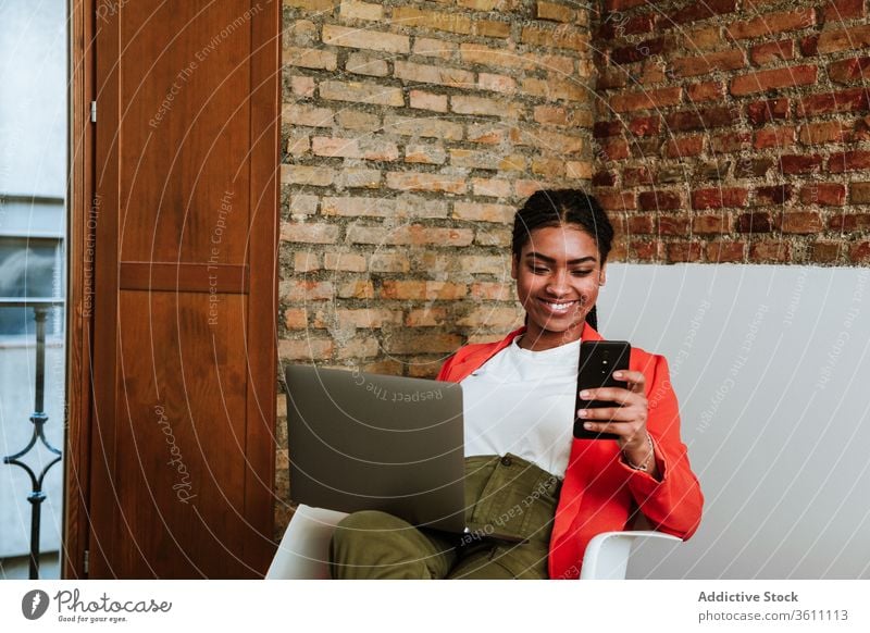 Lächelnde moderne schwarze Frau mit Gadgets im Sessel Business Laptop heiter Smartphone online elegant Computer Loft Design Innenbereich freiberuflich Glück