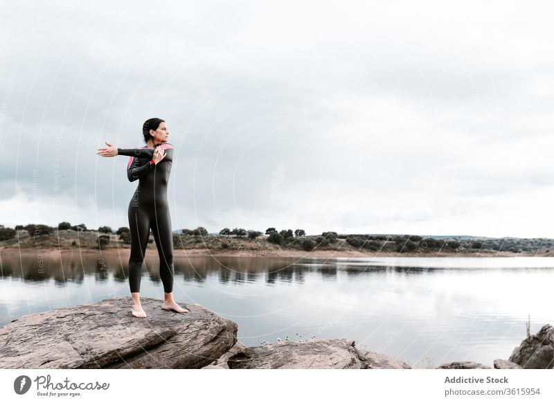 Ruhige Frau streckt sich auf Felsen am Seeufer Dehnung Natur Aufwärmen Taucher Neoprenanzug Harmonie Windstille bedeckt Wassersport Sport Wellness beweglich