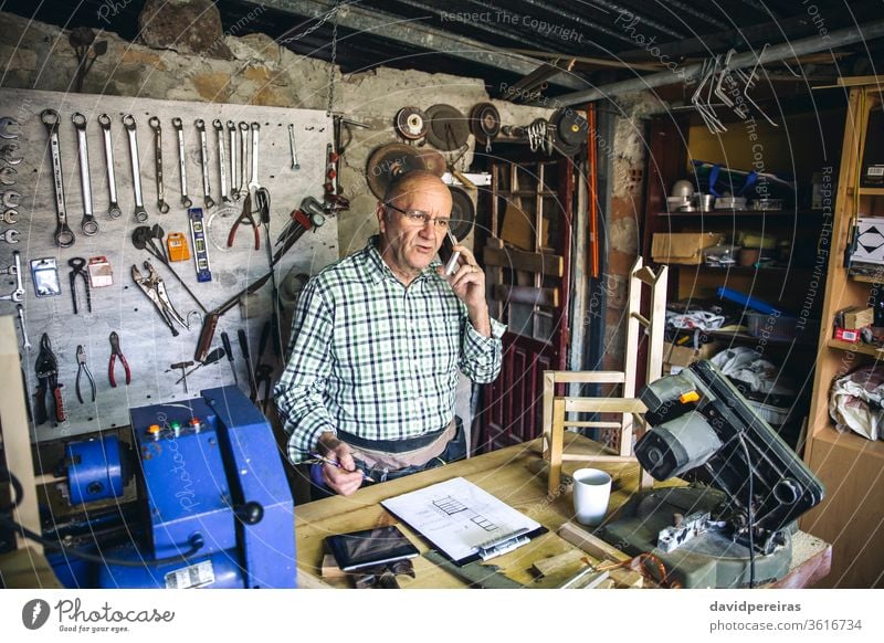 Schreiner in seiner Werkstatt Schreinerei Zimmerer Mann Mobile Senior sprechend Business maßgefertigt Werkbank reif männlich schreibend Holz Arbeiter plant