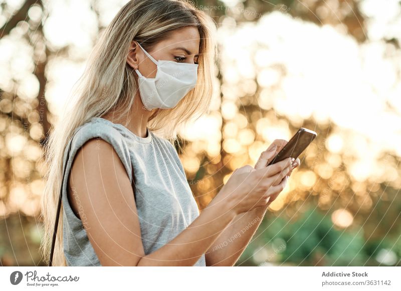 Frau mit Gesichtsmaske auf Smartphone im Park stehend heiter benutzend Glück genießen Browsen trendy Lifestyle Apparatur Gerät soziale Netzwerke online Internet