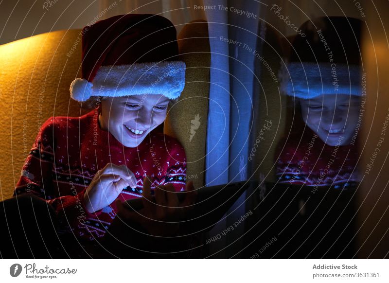 Glückliches Kind genießt neue Tablette am Weihnachtsabend zu Hause Junge Weihnachten Vorabend Weihnachtsmütze Feiertag Zahnfarbenes Lächeln Internet online