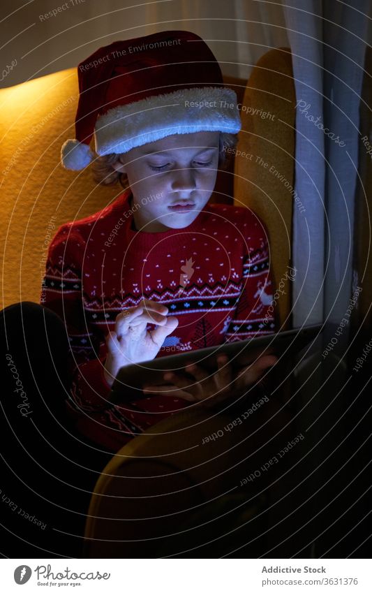 Kind genießt neue Tablette am Weihnachtsabend zu Hause Junge Weihnachten Vorabend Weihnachtsmütze Feiertag Internet online benutzend Apparatur Gerät