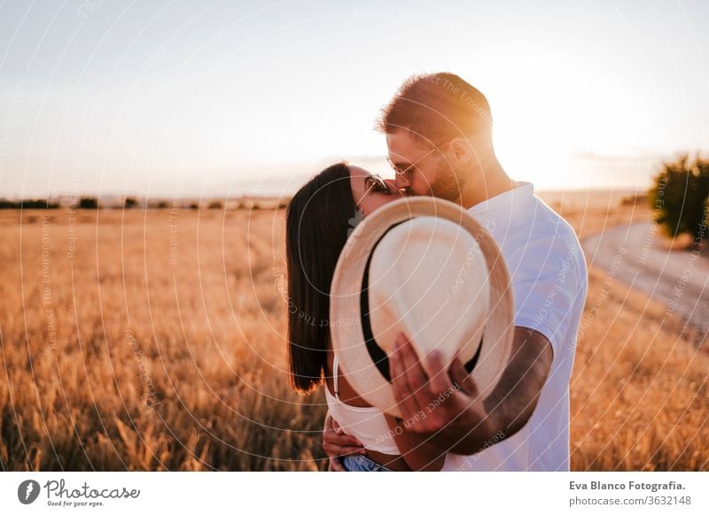 junges Paar, das sich bei Sonnenuntergang in einem gelben Feld küsst und mit einem Hut versteckt. Liebe und Sommerzeit Deckung Tierhaut gelbes Feld Mann Frau