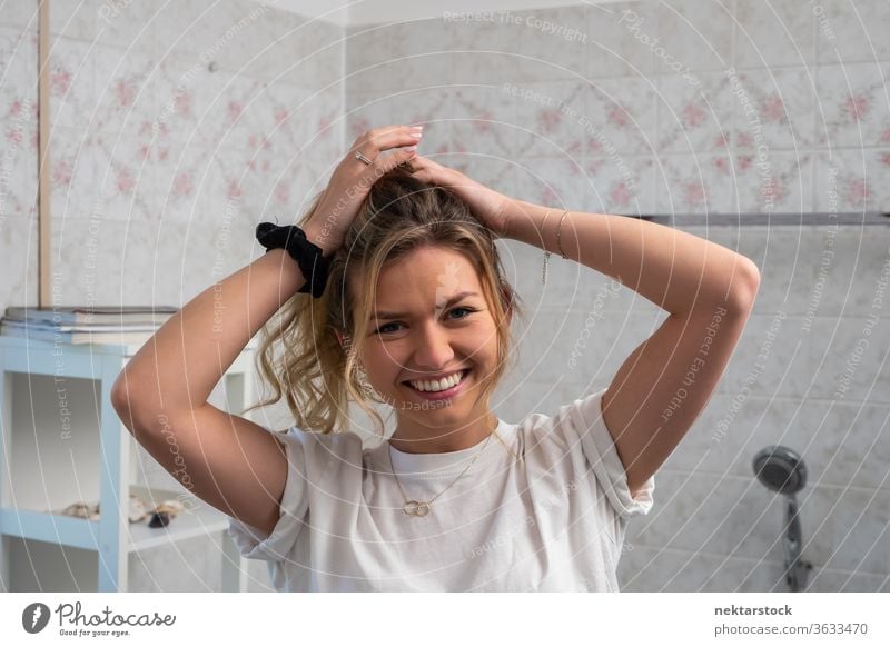 Junge Frau bindet sich die Haare zurück und lächelt im Badezimmer lange Haare Frisur Porträt Lächeln Lifestyle in die Kamera schauen blondes Haar Nur eine Frau