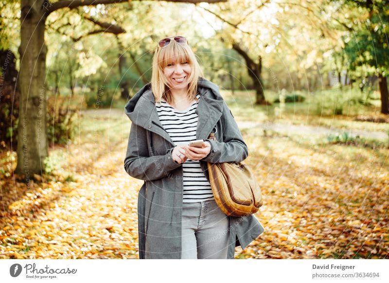 Glückliche blonde Frau in einem herbstlichen Wald oder Park, die mit ihrem Handy SMS schreibt. Kommunikation, Technik und Outdoor-Konzept. Herbst schön Mobile