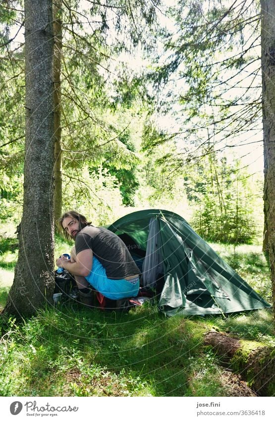Mann sitzt im Wald vor einem Zelt und schaut nach hinten in die Kamera. Junger Mann Morgen Sommer Sonnenlicht Natur Abenteuer Ferien & Urlaub & Reisen