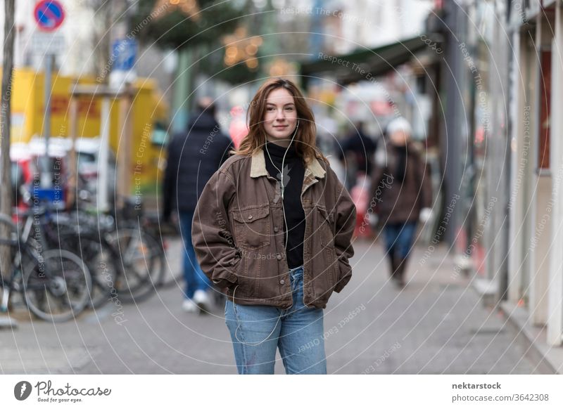 Junge Frau in Jeans und Jacke hört Kopfhörer auf dem Bürgersteig kaukasische Ethnizität Audio Musik hören Straße Freizeitkleidung Lächeln Hände in den Taschen