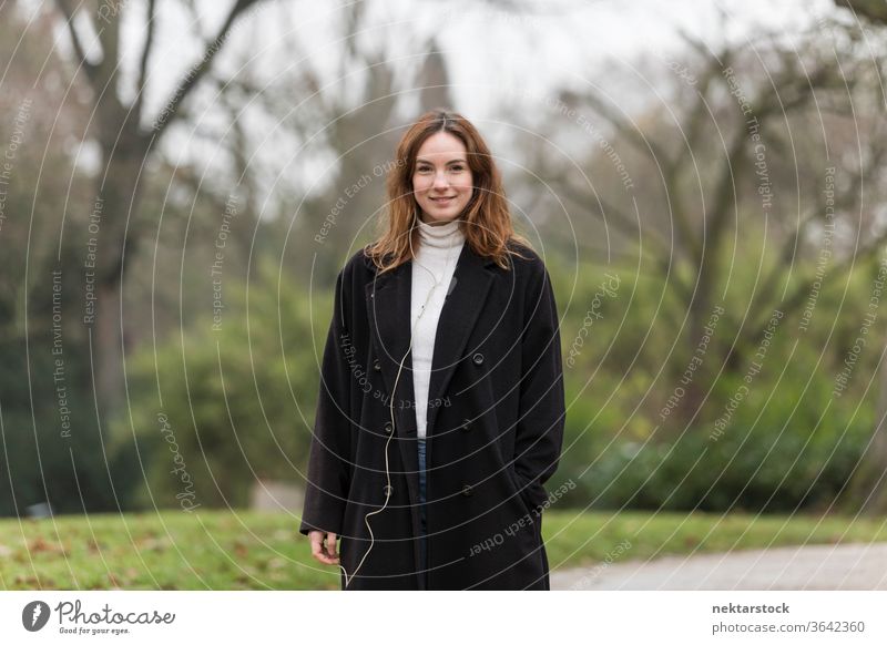 Junge Frau mit braunen Haaren und schwarzem Mantel lächelt im Park kaukasische Ethnizität Kopfhörer Audio Musik hören braune Haare