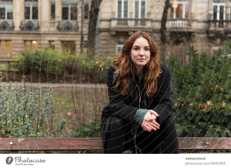 Junge Frau schaut in die Kamera, die auf einer Bank sitzt kaukasische Ethnizität Kopfhörer Audio Musik hören braune Haare Modell aus dem wirklichen Leben