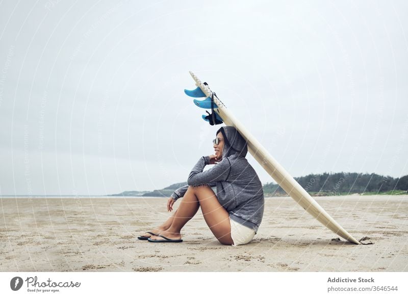 Entspannte Frau sitzt am Strand mit einem Surfbrett auf dem Kopf Surfer ruhen lässig Sand sich[Akk] entspannen sportlich ruhig Sonnenbrille Lifestyle Natur