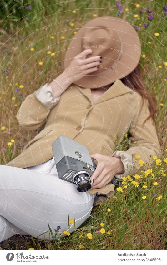 Gesichtslose, auf Gras ruhende Frau mit Retro-Videokamera Fotokamera Hut elegant Wiese Deckblatt Blume Landschaft altehrwürdig Lügen Tierhaut Fotograf retro