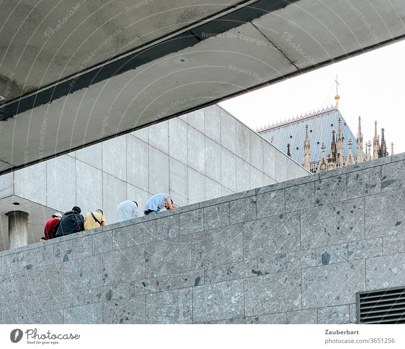 Jugendliche sitzen gebeugt auf einer Mauer aus Steinplatten in moderner Architektur, im Hintergrund ein kleiner Teil des Kölner Doms Wand Linien Stadt