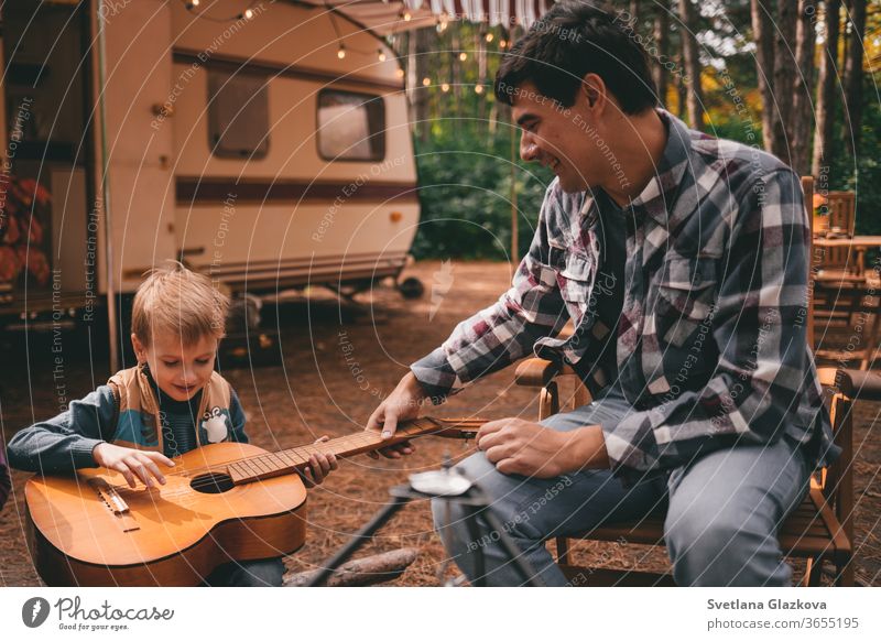 Vater lehrt den Sohn Gitarre spielen, während er sich auf einem Campingausflug im Herbstwald entspannt. Wohnwagen. Herbstsaison Ausflug ins Freie Familie fallen