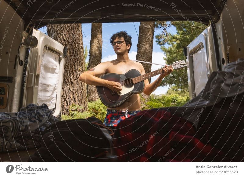 Typ, der im Wald (Camping) Abenteuergitarre spielt, vom Inneren eines Minivans aus gesehen Erholung Gitarre Tourismus Reise Lager Ausflug außerhalb jung Lächeln