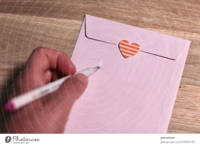Linkshänder. Eine Hand hält einen Stift in Schreibhaltung. Ein Briefkuvert verschlossen mit einem Herzaufkleber wird adressiert. Liebesbrief. Post briefkuvert