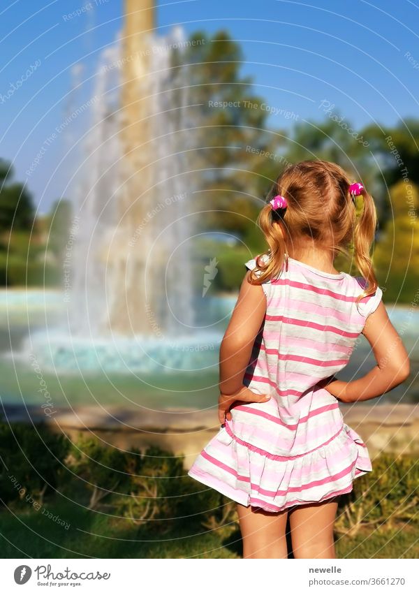Kleines Mädchen im Vorschulalter, das den Brunnen von hinten betrachtet. Kleines weibliches Kind genießt es, im Sommer draußen zu sein. Neugierige Blondine, die auf plätschernde Tröpfchen schaut. Freude an den Ferien. Hübsches Kind Fotografie