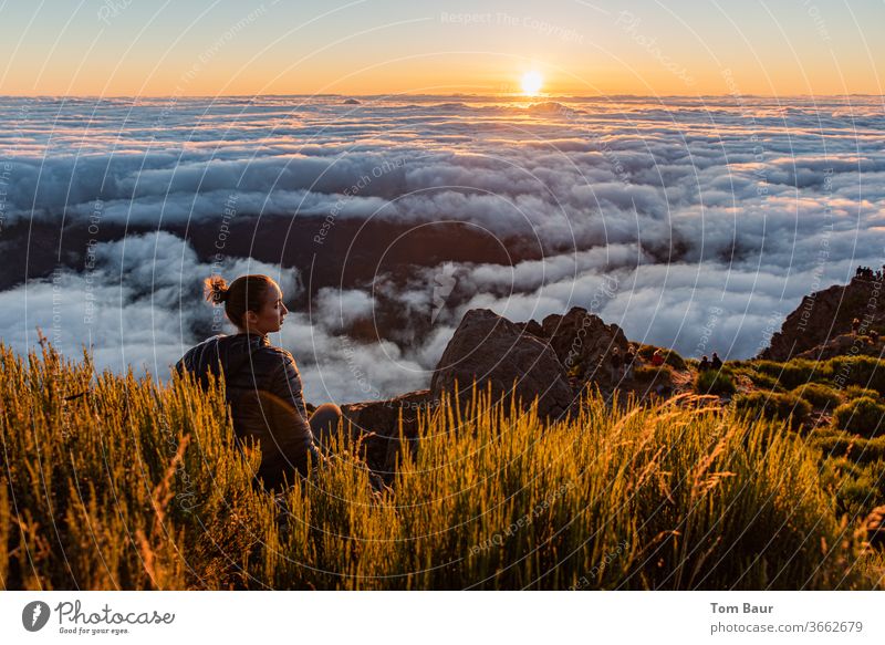 junge Frau am Rand eines Berggipfels sitzt über den Wolken und schaut sich den Sonnenaufgang an. Die aufgehende Sonne am Horizont dicht über einem dichten Wolkenteppich