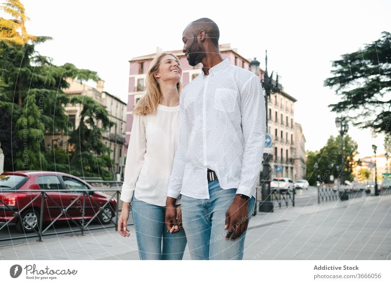 Fröhliches, verschiedenartiges Paar schlendert auf der Straße Spaziergang urban Bürgersteig Händchenhalten Lächeln Glück heiter Großstadt Partnerschaft Romantik