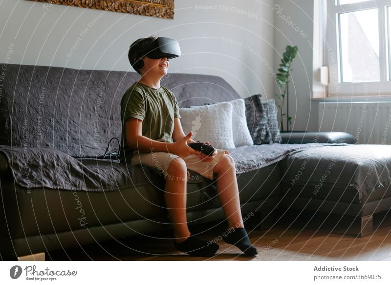 Kind in VR-Headset spielt auf der Couch Junge Schutzbrille spielen Gamepad virtuell Realität benutzend Sofa unterhalten modern heimwärts Gerät wenig bezaubernd