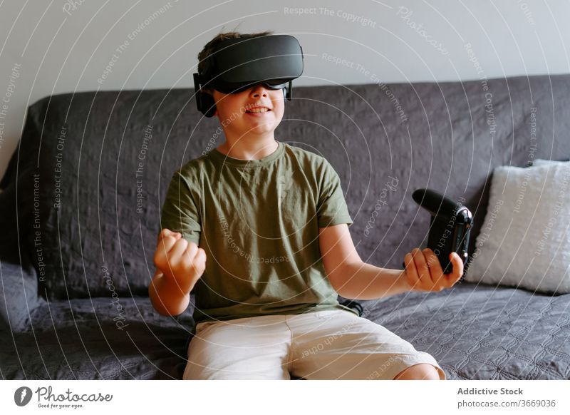 Kind in VR-Headset spielt auf der Couch Junge Schutzbrille spielen Gamepad virtuell Realität benutzend Sofa unterhalten modern heimwärts Gerät wenig bezaubernd