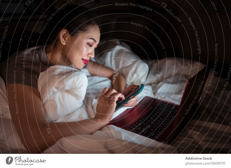 Positive asiatische Frau liegt mit Laptop und Smartphone im Bett sich[Akk] entspannen Internet Komfort heimisch heiter Nacht benutzend Gerät Apparatur Netbook
