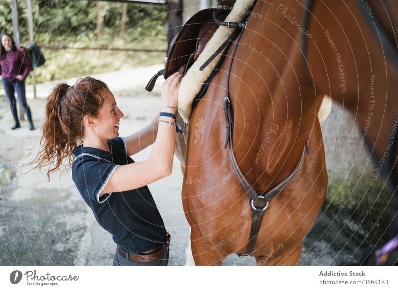 Junge Frau mit Pferd im Stall Reiterin Pferdestall Sattel vorbereiten Tier Dressur Jockey angezogen Zügel Gerät pferdeähnlich jung Konzentration ernst Kastanie