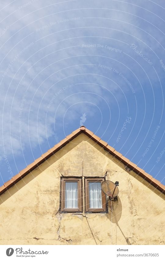 rissige ältere Giebelwand mit zwei kleinen Fenstern und einer Satellitenschüssel mit Schatten vor blauem Himmel mit Schönwetterwolken spitz wohnen Riss