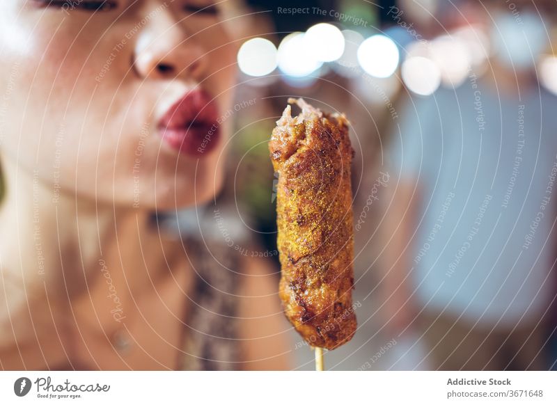 Ethnische Frau bläst auf gebratene Wurst auf der Straße Straßenessen Nacht Markt Lebensmittel Wurstwaren Schlag cool heiß Asiatische Küche asiatisch ethnisch