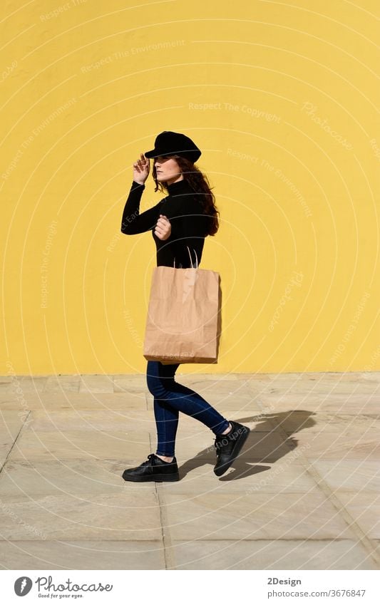 Modeporträt einer Frau mit Einkaufstaschen in schwarzem Outfit mit Hut an gelber Wand posierend. kaufen Tasche Mädchen Sale Glück schön Erwachsener Porträt