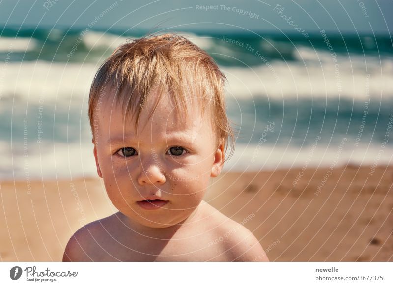 Bezauberndes Kleinkind, das zur Sommerzeit in die Kamera schaut. Junges kaukasisches Baby am tropischen Strand mit der Ozeanwelle hinter ihm. Männliches Kinderporträt bei seinem ersten Urlaub am Sandstrand. Ausdruck von Neugierde.