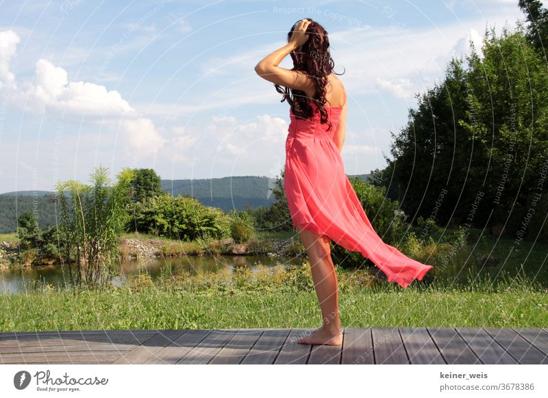 Eine junge Frau in wehendem roten Kleid steht barfuß auf einer Terrasse vor grüner Landschaft mit kleinem See langhaarig Locken Barfuß Garten Urlaub Natur Wald