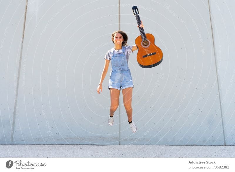 Optimistischer Teenager mit Gitarre springend Musiker Lächeln Wand Gebäude lässig Sommer Energie Hobby Mädchen Glück akustisch modern Großstadt Straße heiter