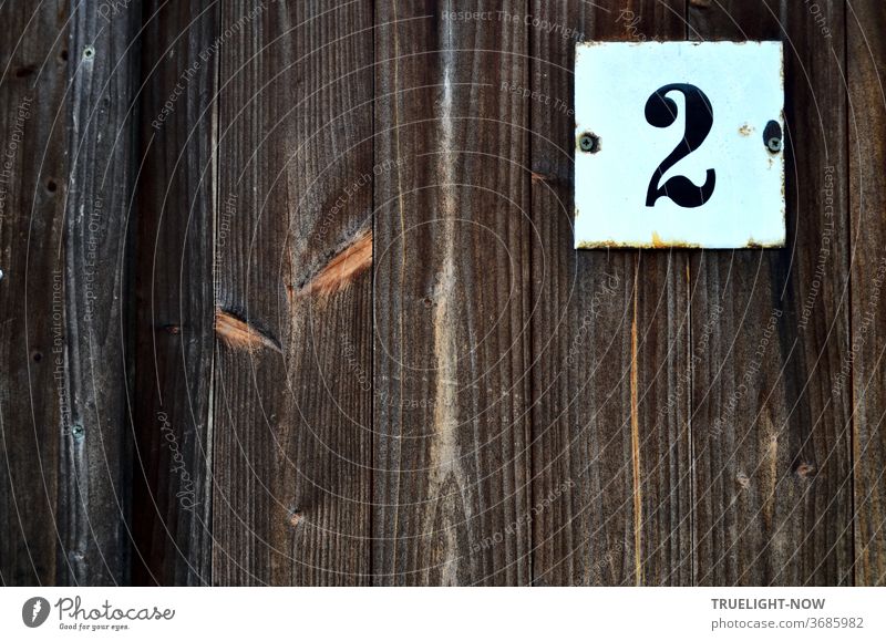An einem alten, Blick dichten Zaun aus Holz Brettern in senkrechter Anordnung ist ein weißes quadratisches  Metall Schild angeschraubt, auf dem eine schwarze, auffällige Ziffer 2 in Antiqua Schrift vermutlich die Hausnummer markiert