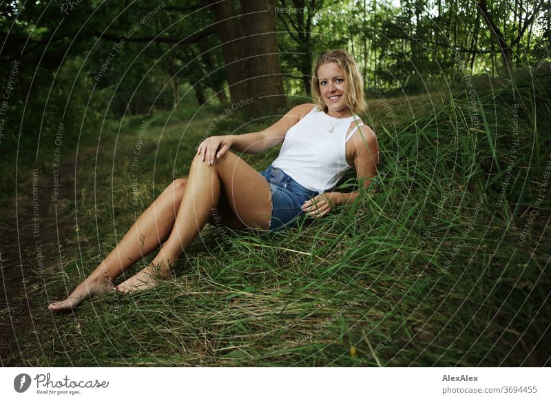 Portrait einer jungen Frau die in der Natur sitzt und lächelt junge Frau blond Lächeln Schmuck schön langhaarig Landschaft gebräunt selbstbewußt Sommer