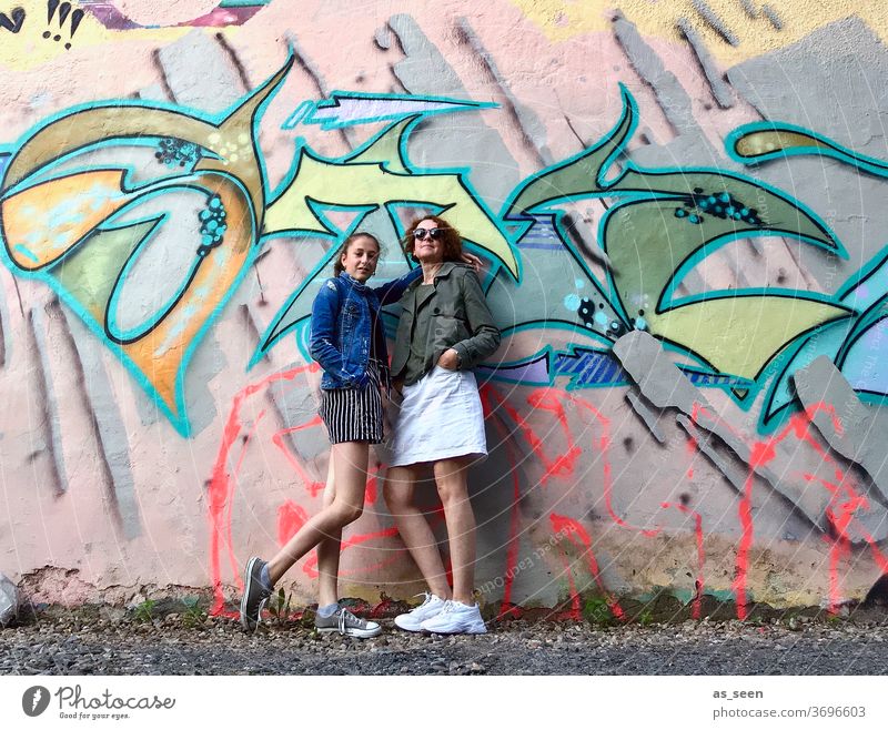 Zwei Frauen vor einer Wand mit Graffiti Sneaker Sonnenbrille gelockt rothaarig Außenaufnahme Tag Mensch Mode grün khakigrün bunt Muster anlehnen cool Coolness
