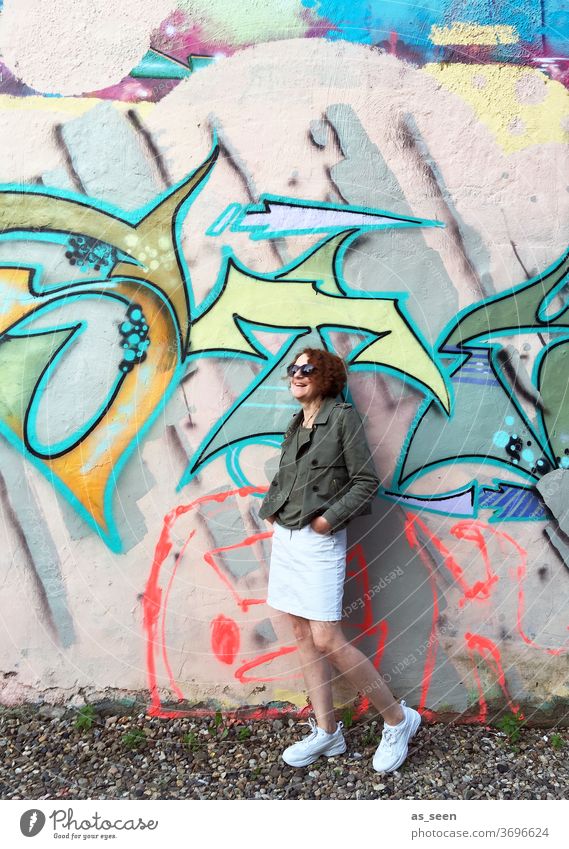 Frau mit Sonnenbrille vor einer Wand mit Graffiti Sneaker gelockt rothaarig Außenaufnahme Tag Mensch Mode grün khakigrün bunt Muster anlehnen cool Junge Frau