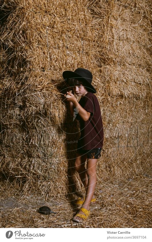 Ein Junge mit Hut, der im Sommer auf den Feldern auf dem Land in einem Strohballenhintergrund spielt Bauernbursche hockend Pflanze Spielen Herbst Kind niedlich