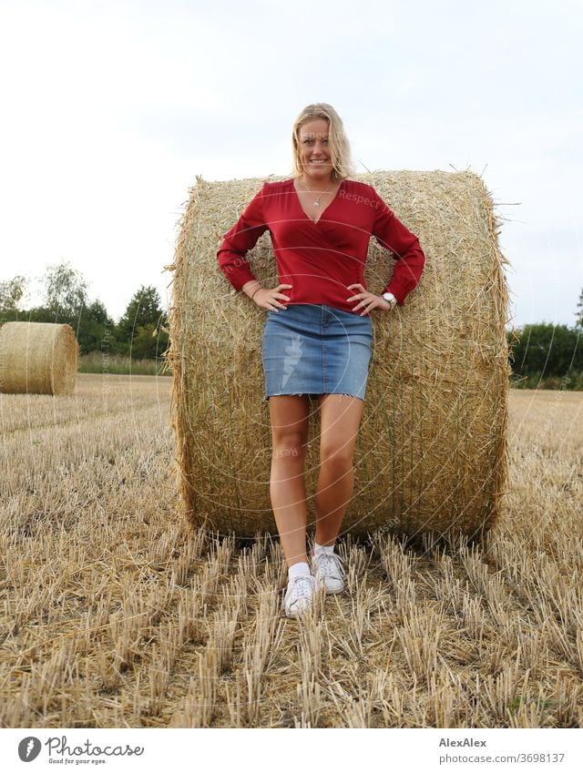 Junge Frau steht an einen Strohballen gelehnt auf dem Feld und lächelt Beine Freizeit Spaß Freude Jeans Ganzkörper Zentralperspektive Blick in die Kamera