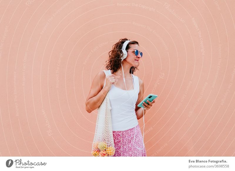 Frau, die ein Mobiltelefon benutzt, Musik über Kopfhörer hört. mit einem Baumwollbeutel mit Obst durch die Stadt läuft. Umweltfreundlich, Null-Abfall-Konzept