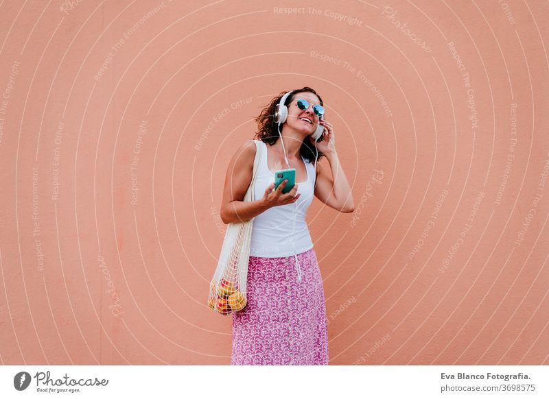 Frau, die ein Mobiltelefon benutzt, Musik über Kopfhörer hört. mit einem Baumwollbeutel mit Obst durch die Stadt läuft. Umweltfreundlich, Null-Abfall-Konzept