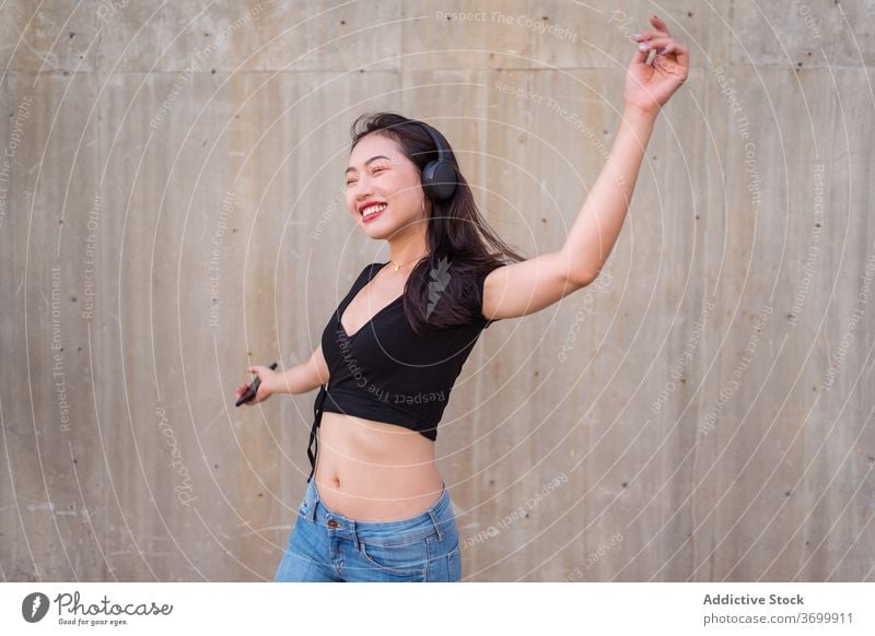 Unbekümmerte Frau, die mit Kopfhörern Musik hört zuhören Tanzen Straße sorgenfrei Smartphone benutzend genießen ethnisch asiatisch Glück heiter Freude Klang