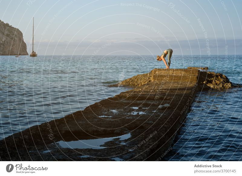 Schlanke Frau steht in Yoga-Pose in der Nähe von Meer MEER üben Asana Vorwärtsbeuge stehen uttanasana Gleichgewicht Windstille Meeresufer Stein felsig Harmonie