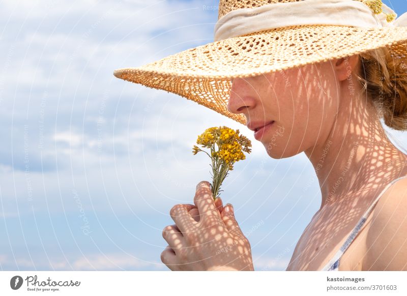 Porträt einer jungen, schönen, fröhlichen Frau mit Stroh-Sonnenhut, die einen kleinen Strauß gelber Wildblumen vor blauem Sommerhimmel riecht Blumenstrauß