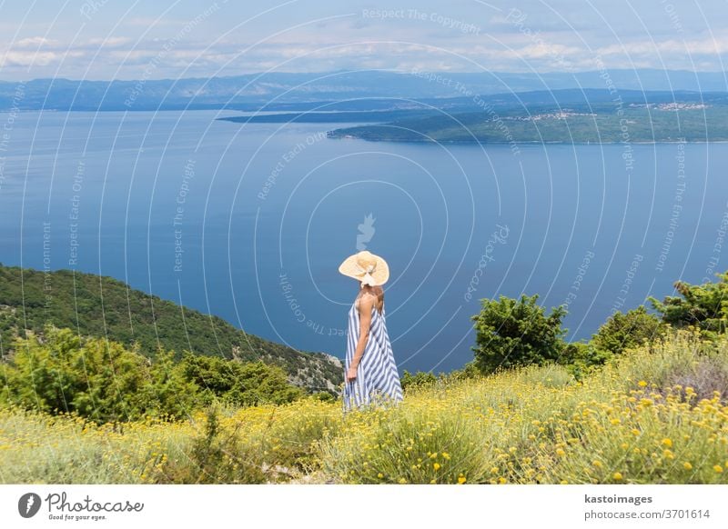 Rückansicht einer jungen Frau in gestreiftem Sommerkleid und Strohhut, die in einer Superblüte von Wildblumen steht und sich entspannt, während sie einen schönen Blick auf die Natur der Adria genießt, Kroatien